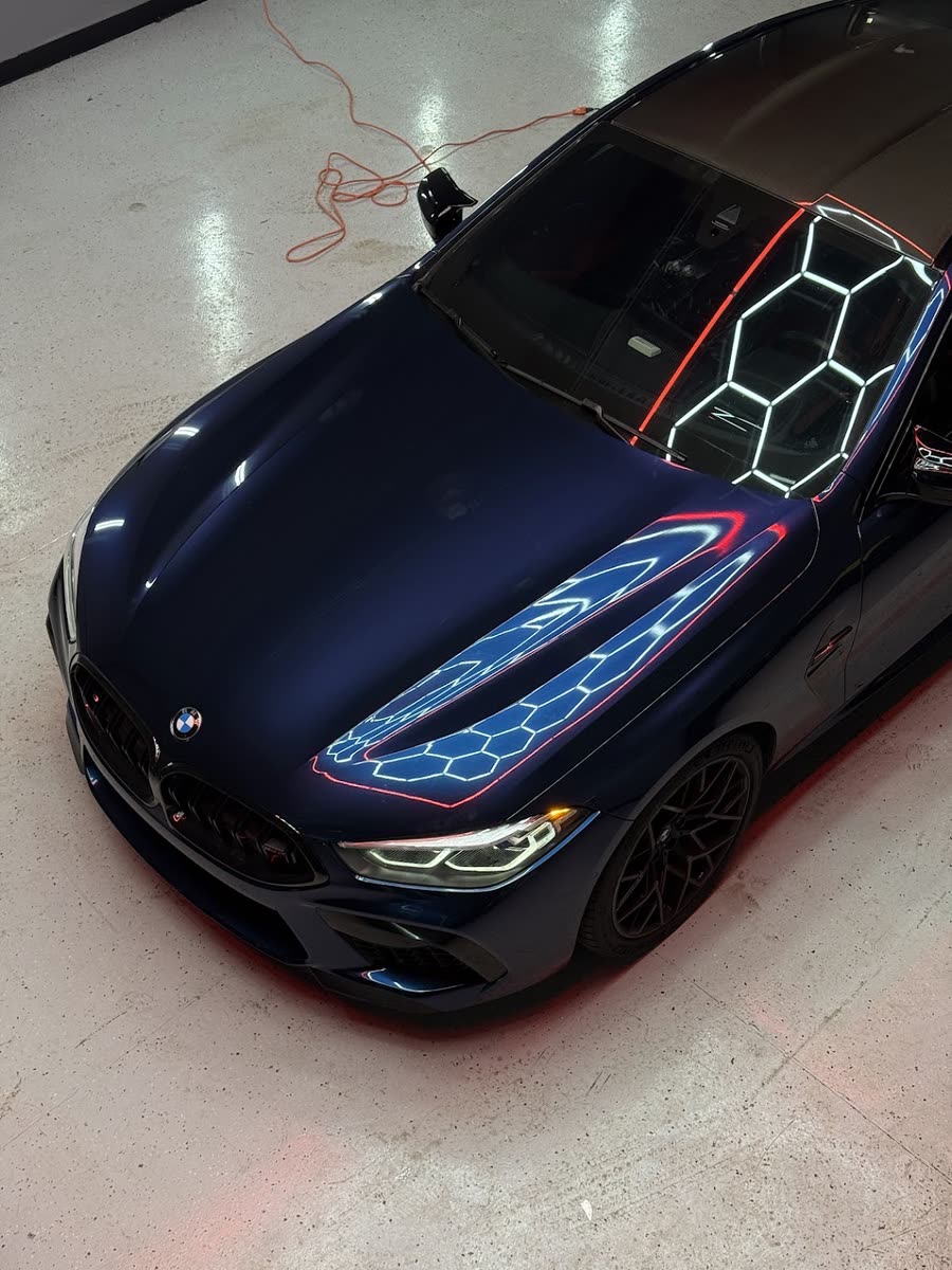 Navy blue BMW M8 seen from above inside the Perfect Graphics shop, hex lighting reflected across the hood and roof