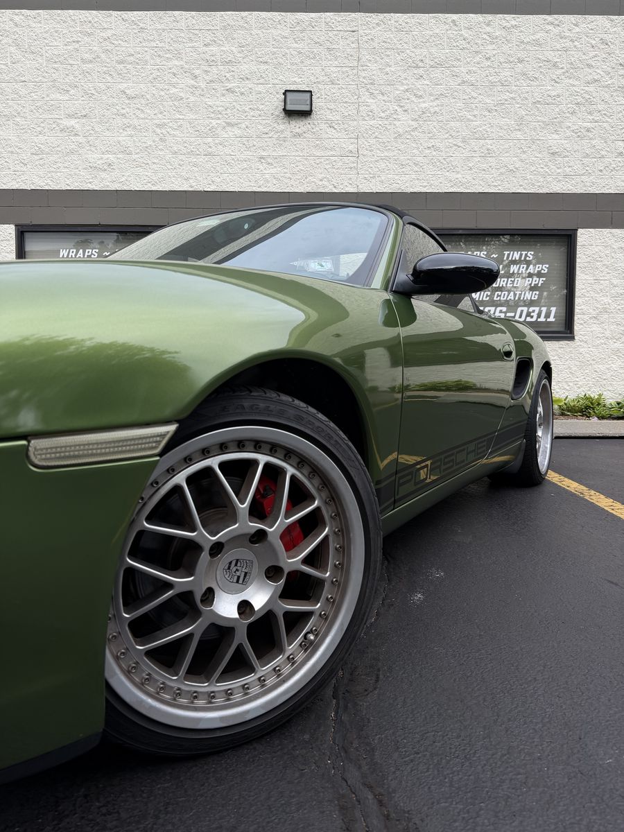 Front wheel and fender of an olive green wrapped Porsche, red brake caliper behind the spokes