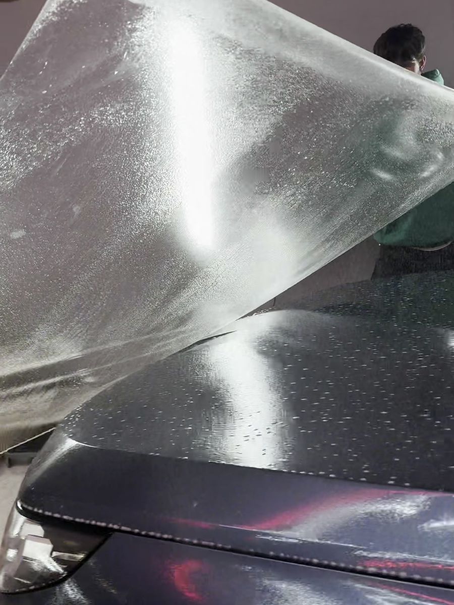 A wet sheet of paint protection film being laid over the hood of a dark car by hand