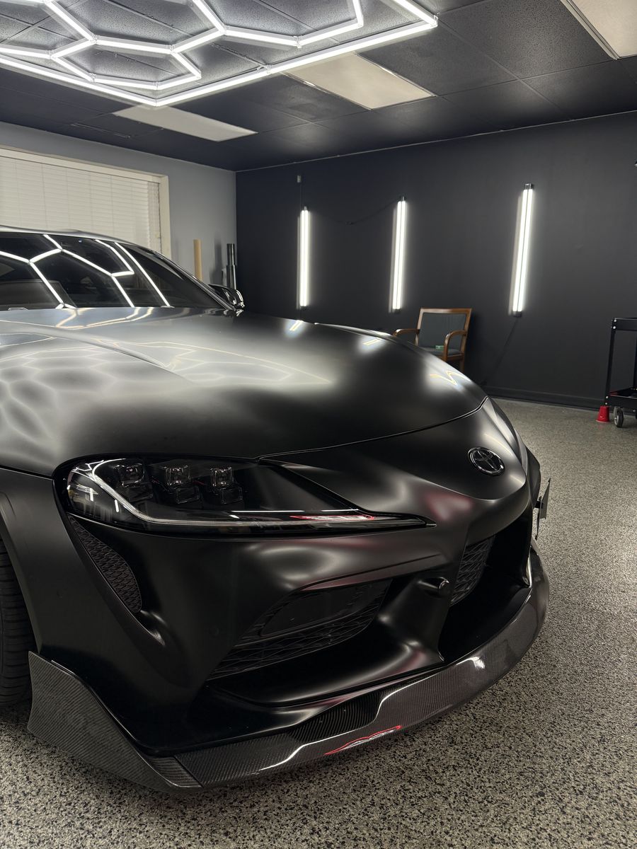Toyota Supra in a satin black wrap, front three-quarter under the hex lighting