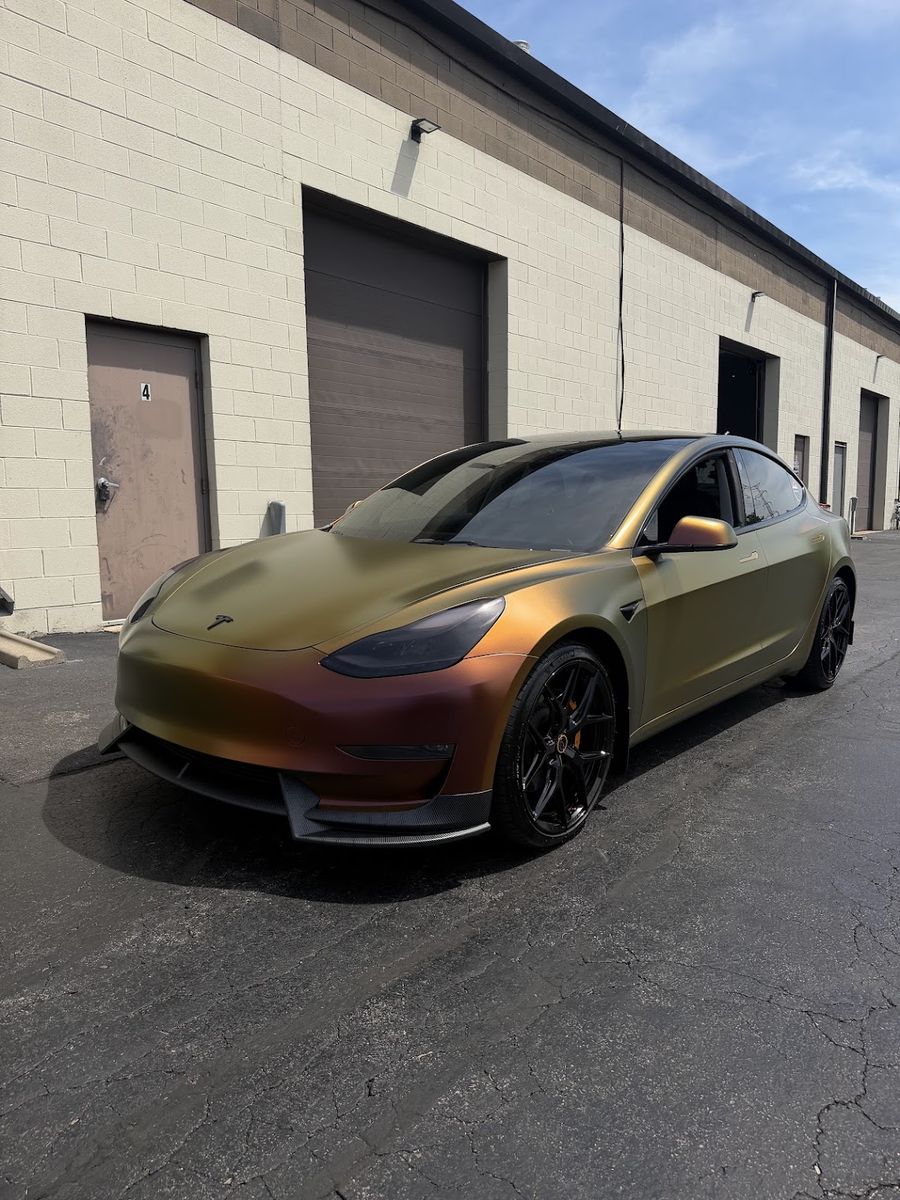 Tesla Model 3 in a gold and bronze color shift wrap, front three-quarter view beside the shop bays